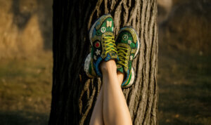 Pernas cruzadas apoiadas num tronco de árvore, usando ténis verdes da Exé decorados com padrões coloridos, flores, símbolos de paz e borboletas, num ambiente natural ao ar livre.